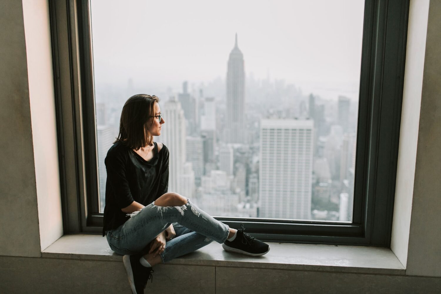 woman in black jacket and blue denim jeans sitting on window with the view on empire state building